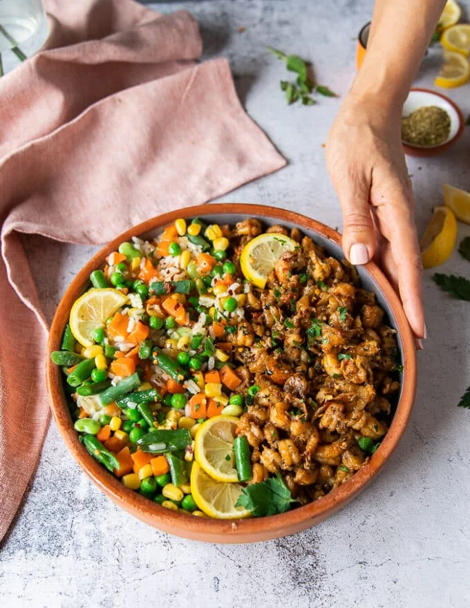A hand holding a plate of cooked crawfish tail and served with rice, surrounded by lemon slices on a marble board