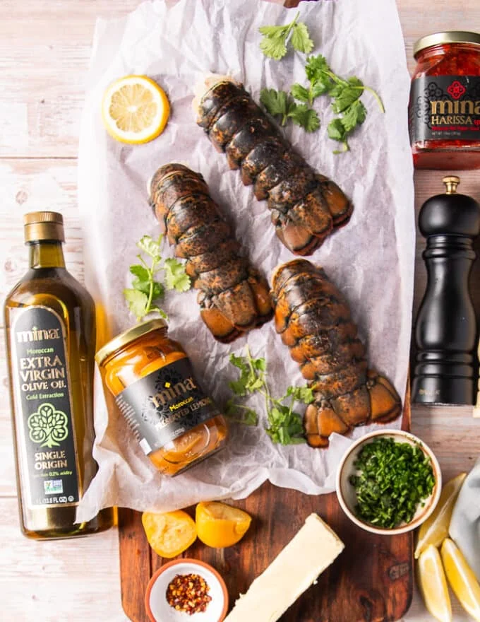 Ingredients to make grilled lobster tail including fresh lobster tails, some preserved lemons, some butter, fresh herbs, some olive oil, salt and pepper