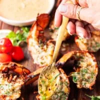 A hand pouring melted butter over the lobster tails off the grill