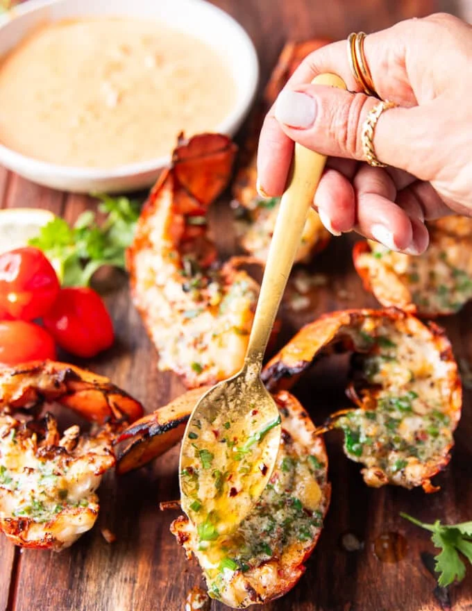 A hand pouring melted butter over the lobster tails off the grill