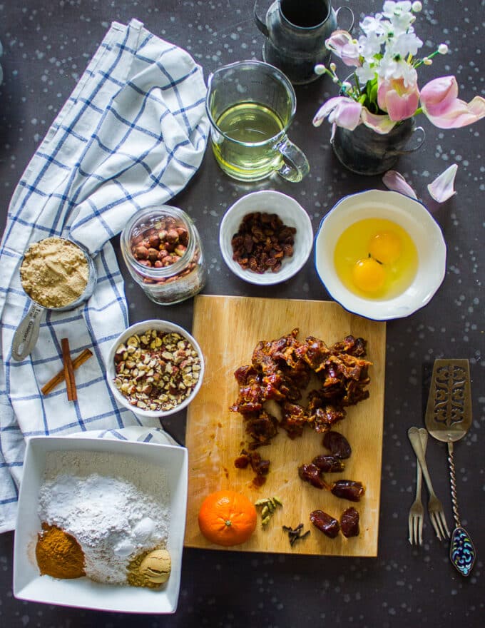 date cake Ingredients on a board including pitted dates, some orange zest and juice, some bornw sugar in a bowl, 2 eggs in a bowl, some chai spice, some ground cinnamon, ground ginger in bowls, some flour in a bowl, baking powder, baking soda in a bowl, and oil in a bowl
