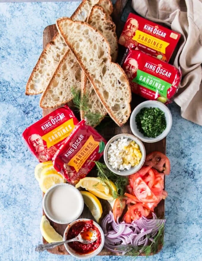 ingredients to make sardines on toast include canned sardines in a tin, some lemon, lemon hot sauce, tahini sauce, sliced tomatoes on a board, sliced onions, some crunchy bread slices, some seasoning and fresh dill