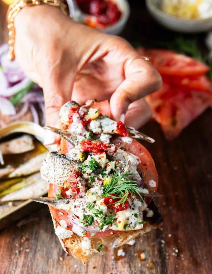 A hand holding a toast of sardines with all the toppings like lemon, feta onions, tomatoes, chilli flakes and more close up showing the texture and layering of the toast