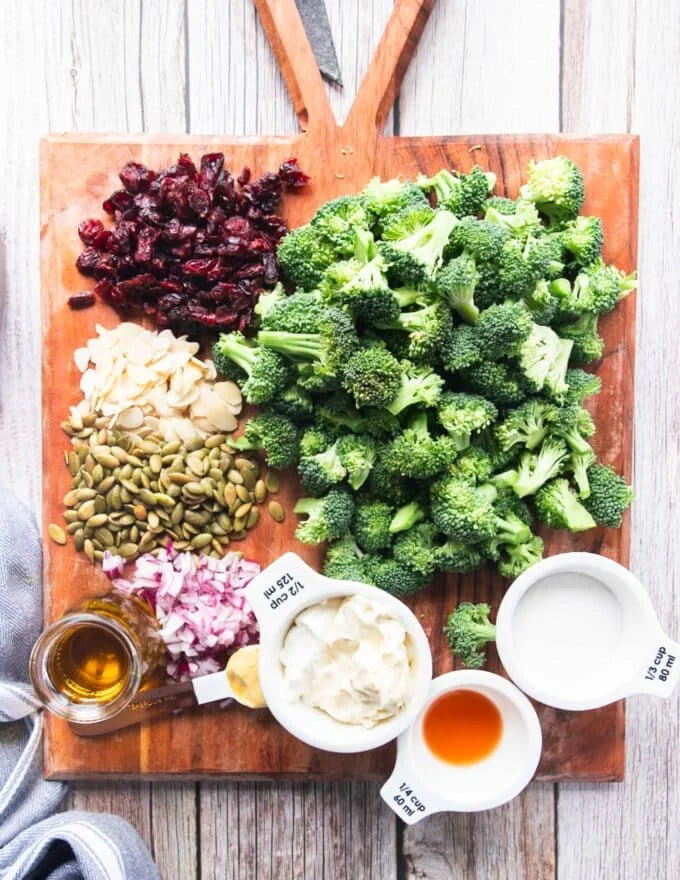 broccoli salad ingredients on a board with some fresh broccoli florets, dried cranberries, seeds, nuts and dressing ingredients