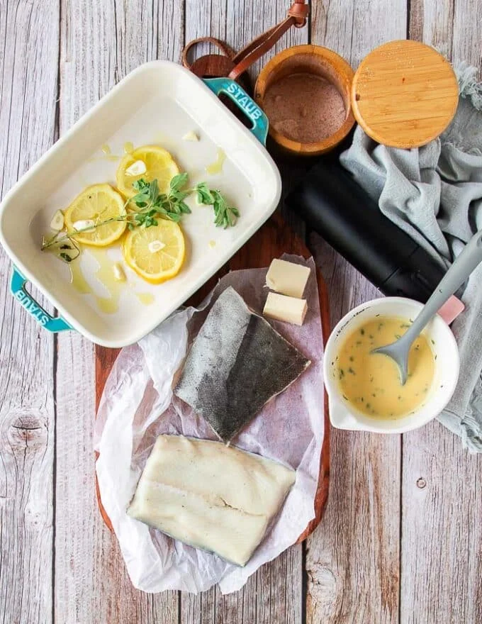 Ingredients for baked chilean sea bass on a board including the fish, butter, lemon, herbs and beurre blanc