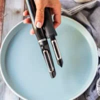A hand holding a vegetable peeler and mandolin to start slicing the beets