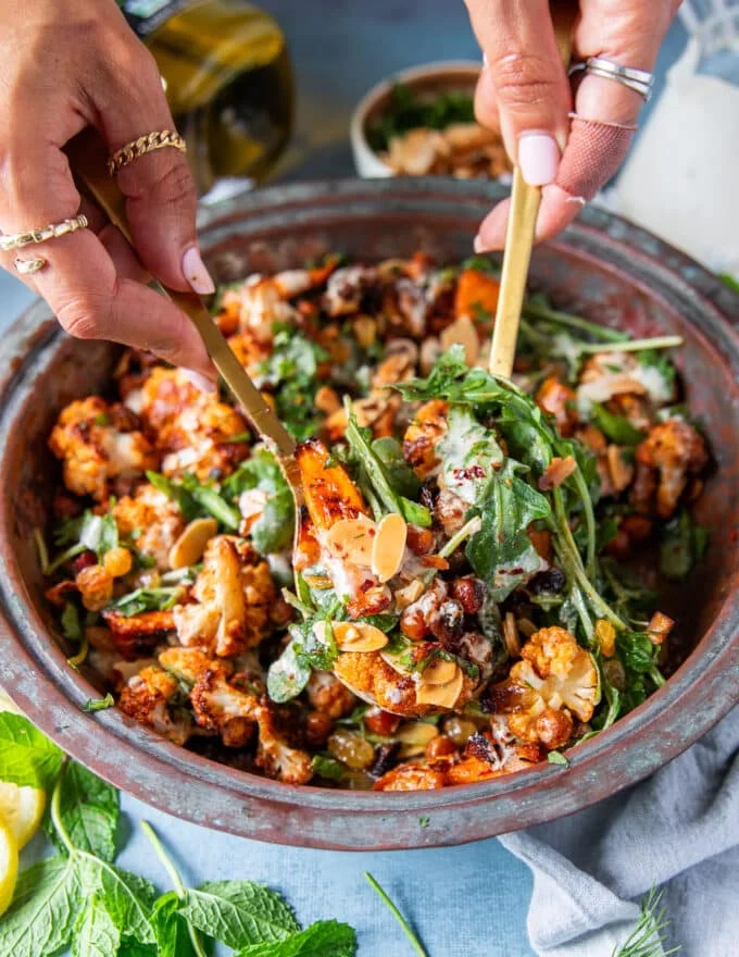 A hand scooping some cauliflower salad with almonds, raisins and chickpeas in tahini dressing using two spoons close up showing the textures
