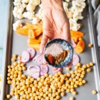 a hand holding a bowl of warm spices to season the cauliflower and chickpeas, onion and carrots before roasting
