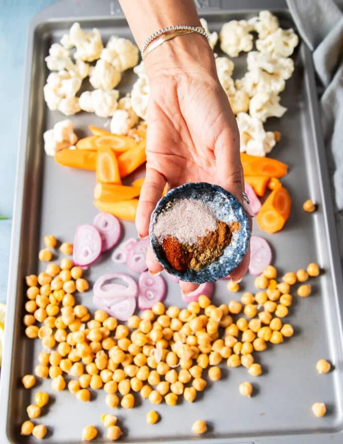 a hand holding a bowl of warm spices to season the cauliflower and chickpeas, onion and carrots before roasting