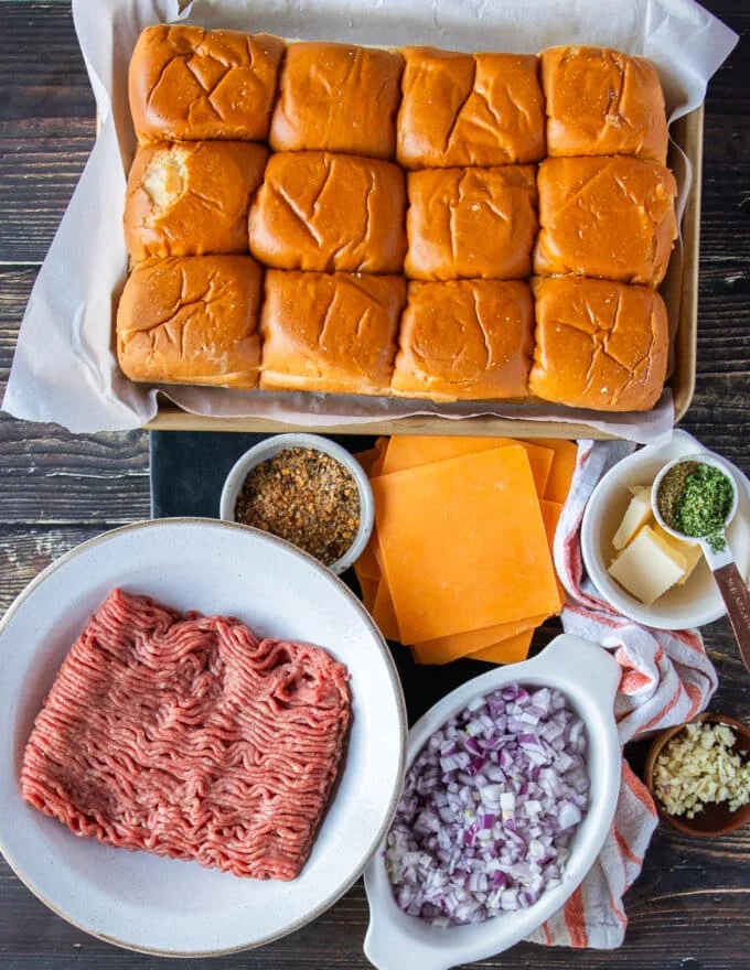 All ingredients needed to make cheeseburger sliders on a board in small bowls with raw beef, bread, onions, seasoning, cheeses, sauces and topping