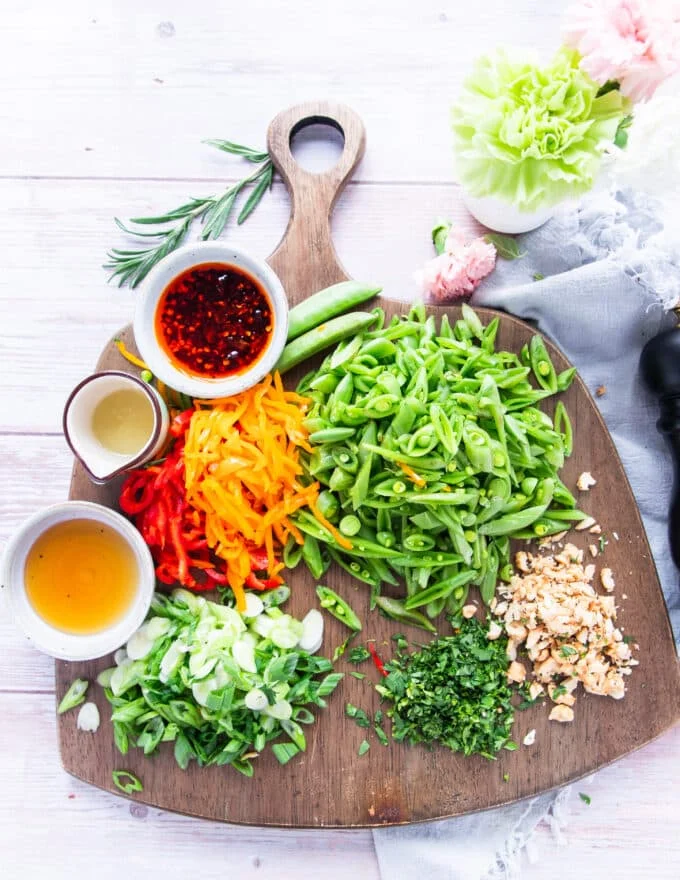 Ingredients for snap pea salad including a variety of fresh veggies and snap peas, raw and chopped, a variety of nuts and small bowls of lime, honey and chilli oil for the dressing