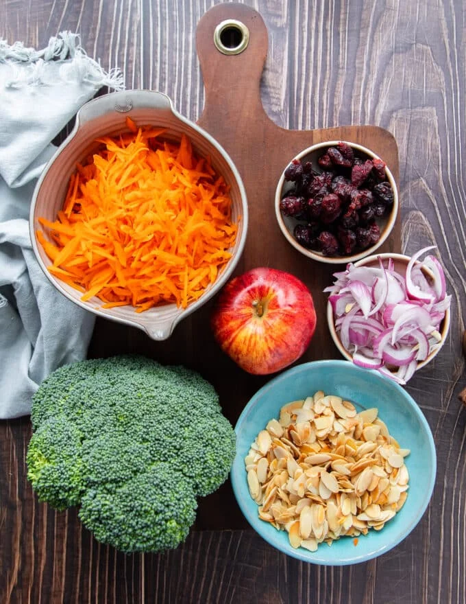 2 heads of broccoli, some shredded carrots, an apple, some sliced onions, a bowl of toasted almonds and a coleslaw dressing in small bowls as ingredients needed to make broccoli slaw