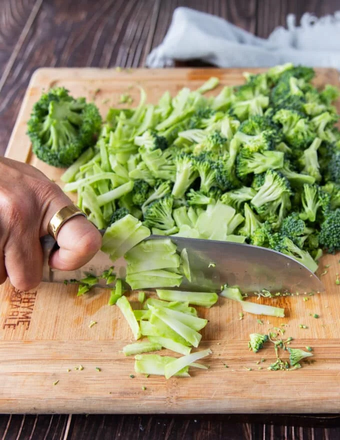 A hand holding a knife to thinly slice the broccoli stalks to make broccoli slaw