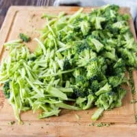Broccoli stalks and florets are shredded thinly on a wooden board ready to make broccoli slaw