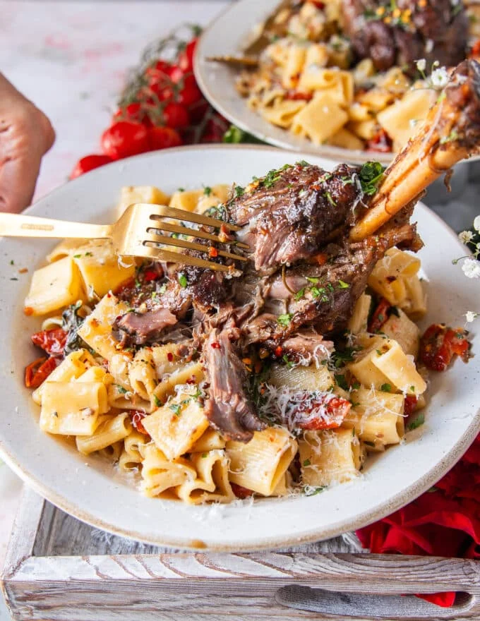 A fork breaking apart a slow cooked lamb shank over pasta showing how fall of the bone it is