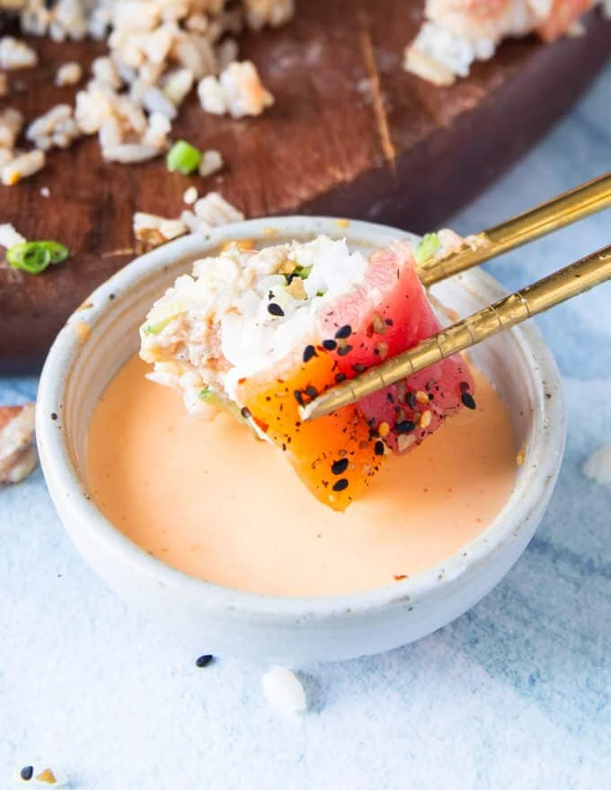 A piece of sushi cake bite is dipped into a bowl of spicy mayo