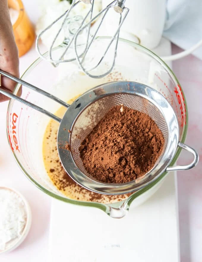 Cocoa powder being sifted over the bowl of light and fluffy eggs and sugar mixture