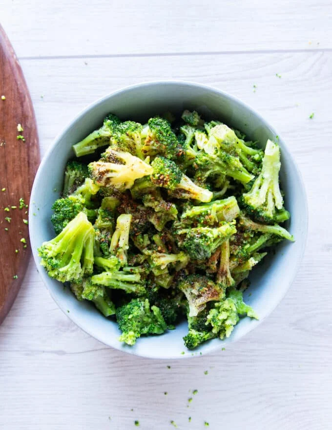 frozen broccoli florets are in a large bowl tossed with seasoning and oil to prepare for air frying
