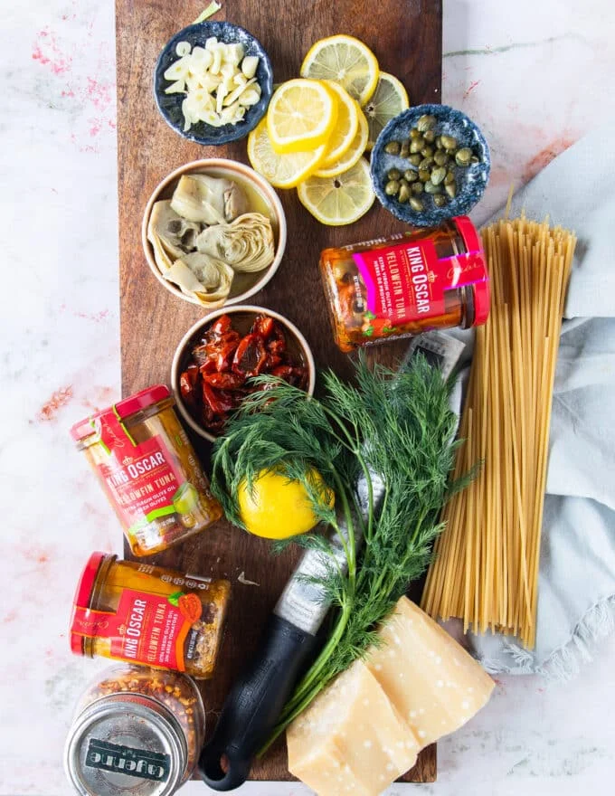 All ingredients needed to make tuna pasta on a board including canned tuna, fresh herbs, pasta, bowls of sun dried tomatoes, bowl of capers, lemon zest, garlic slices, seasoning and parmesan block