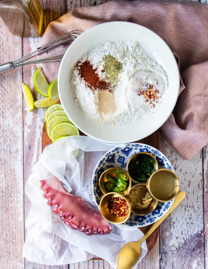 Ingredients needed to make fried octopus including boiled octopus, flour and seasoning in small bowls, some lemon juice, garlic, chilies, olive oil and fresh herbs, and oil for shallow frying