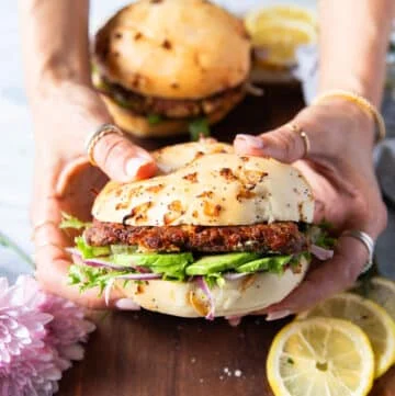 Two hands holding a tuna burger close up showing the layers of avocado, bun, tuna burger, lettuce and other toppings