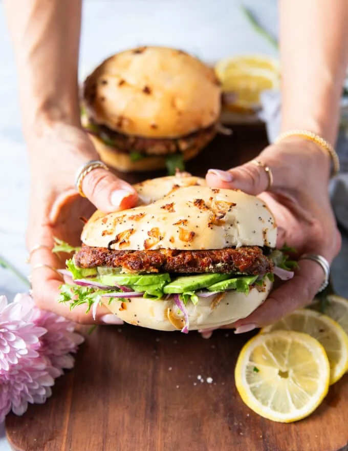 Two hands holding a tuna burger close up showing the layers of avocado, bun, tuna burger, lettuce and other toppings