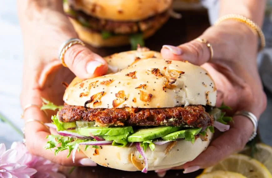 Two hands holding a tuna burger close up showing the layers of avocado, bun, tuna burger, lettuce and other toppings