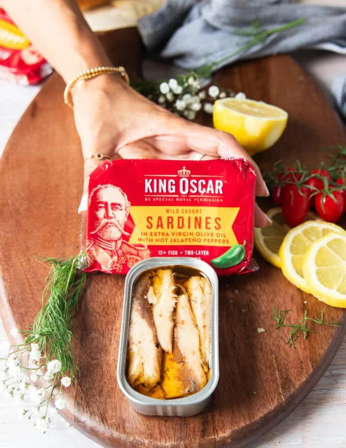 A hand holding two cans of sardines and one canned sardine open showing how it looks inside