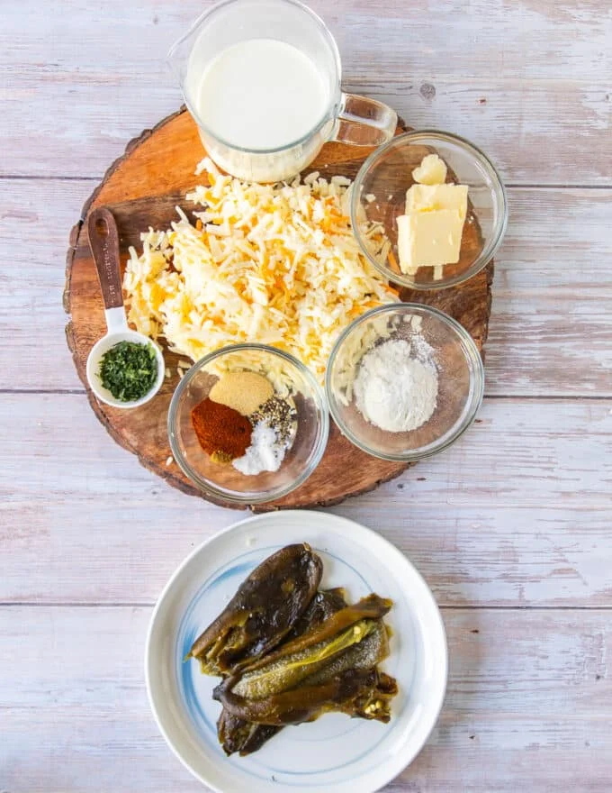ingredients to make smoked queso on a board including cheeses, seasoning, herbs, roasted poblanos, milk, butter and flour