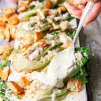 A hand drizzling some creamy caesar dressing over the grilled romaine over a serving board