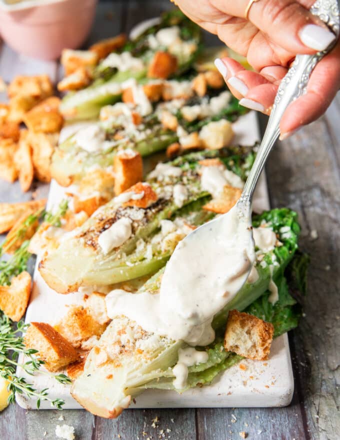 A hand drizzling some creamy caesar dressing over the grilled romaine over a serving board