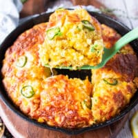 A spatula serving up a spoonful of jalapeno cheddar cornbread from the cast iron skillet showing the texture and studded corn and jalapeño slices on top with a crisp cheese top