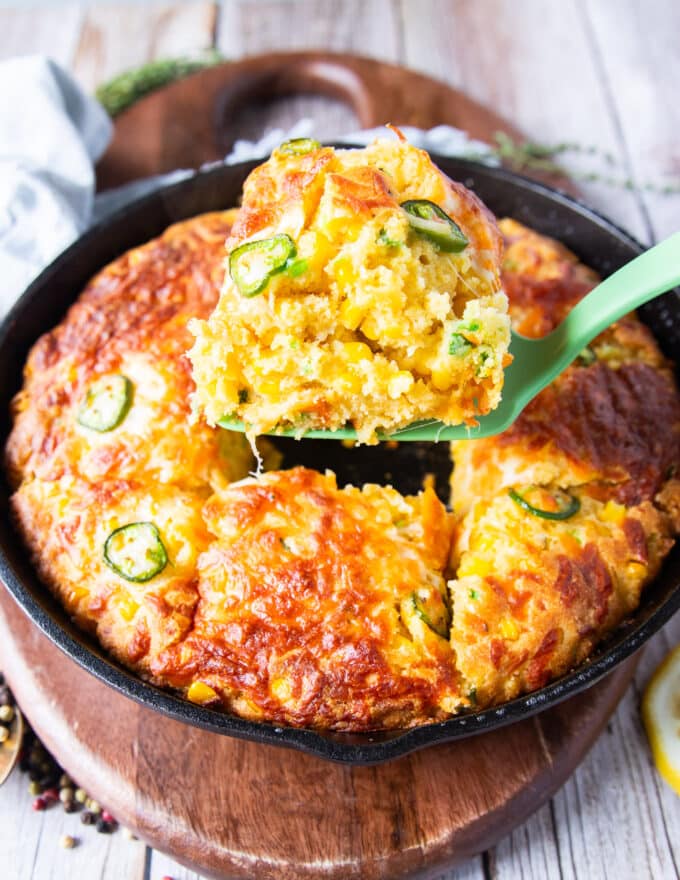 A spatula serving up a spoonful of jalapeno cheddar cornbread from the cast iron skillet showing the texture and studded corn and jalapeño slices on top with a crisp cheese top