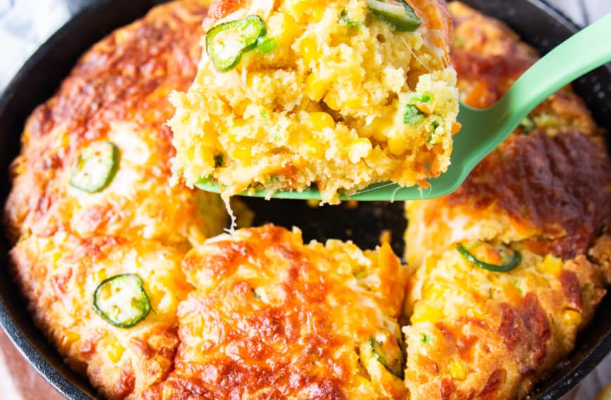 A spatula serving up a spoonful of jalapeno cheddar cornbread from the cast iron skillet showing the texture and studded corn and jalapeรฑo slices on top with a crisp cheese top