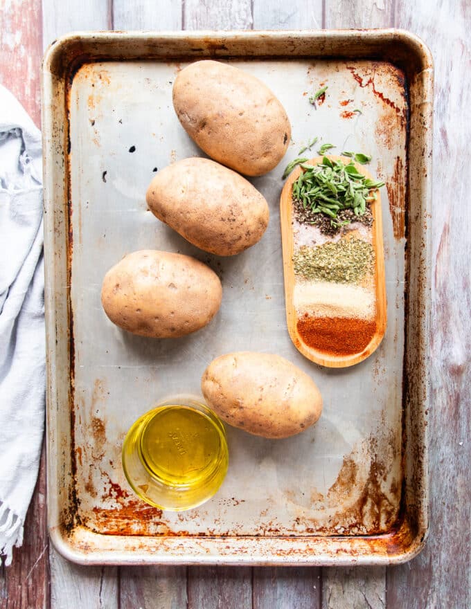 ingredients needed to make potato wedges in a large baking sheet with whole large russet potatoes and a plate of seasoning and a bowl of oil
