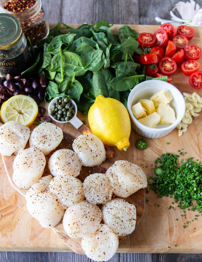 Ingredients on a board to make the scallop pasta including large scallops, spinach, tomatoes, garlic, capers, lemon zest, olive oil and herbs